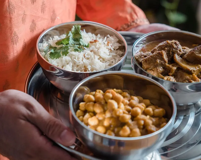 Hands holding steel bowls of home-cooked rice, chana and sabzi, a balanced Indian meal